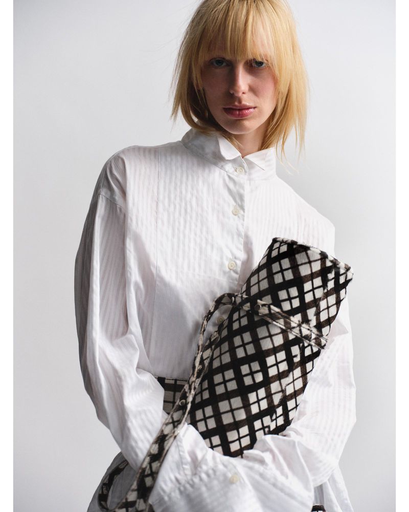 Fashion portrait of a woman in a white shirt holding a checkered bag against a white background in a studio setting with neutral colors.