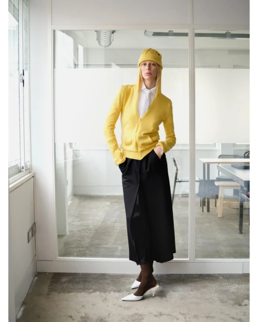 A woman in a yellow hat and jacket stands indoors, showcasing a stylish outfit with a white shirt, black skirt, and white heels.