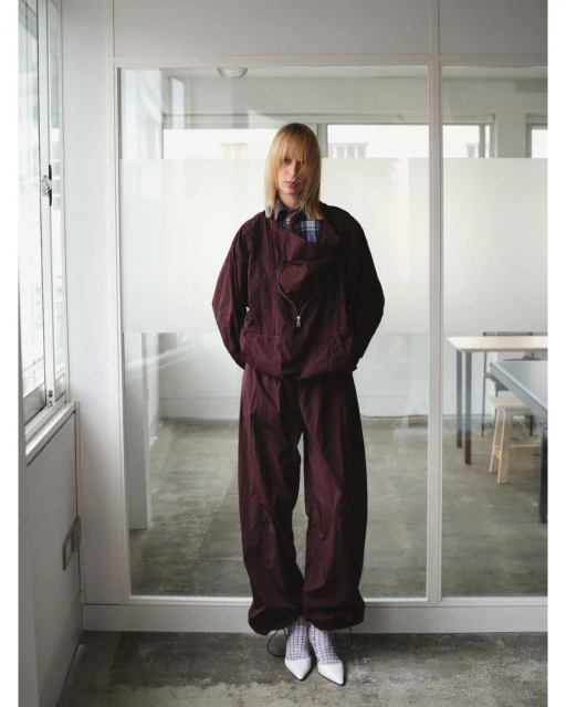 A full-length portrait of a woman in a maroon tracksuit, patterned heels, posing indoors by a window, with a modern office visible.