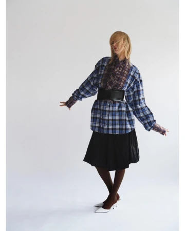 Model in layered plaid shirt and black skirt with wide belt against a white backdrop