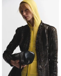 Model wearing a yellow hooded top under a dark tweed jacket holding a black cylindrical bag against a white seamless background