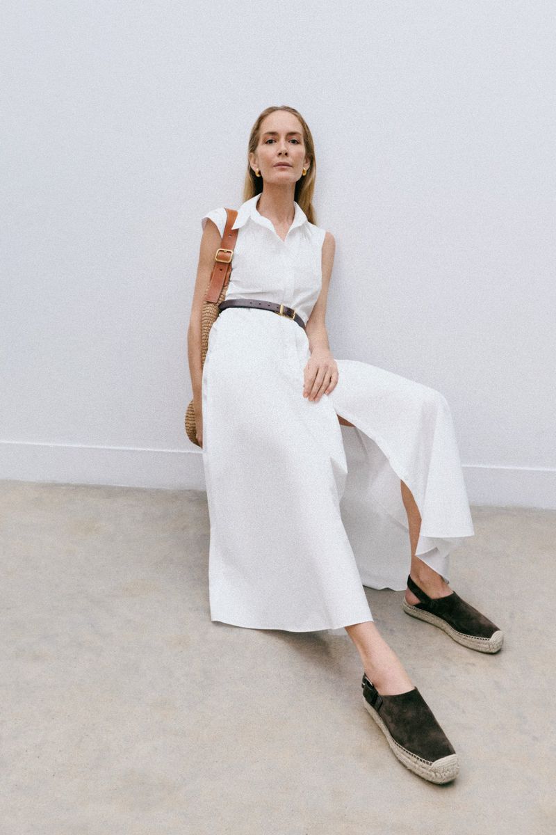 Blonde woman poses while seated in a white dress with a belt, sandals, and straw bag against a white wall in a studio setting.