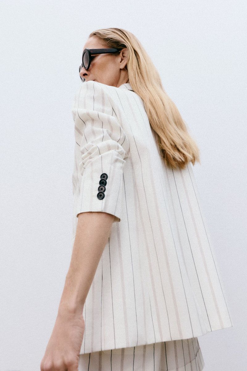 Woman in a striped blazer and sunglasses looks back over her shoulder in a fashion pose against a white background.