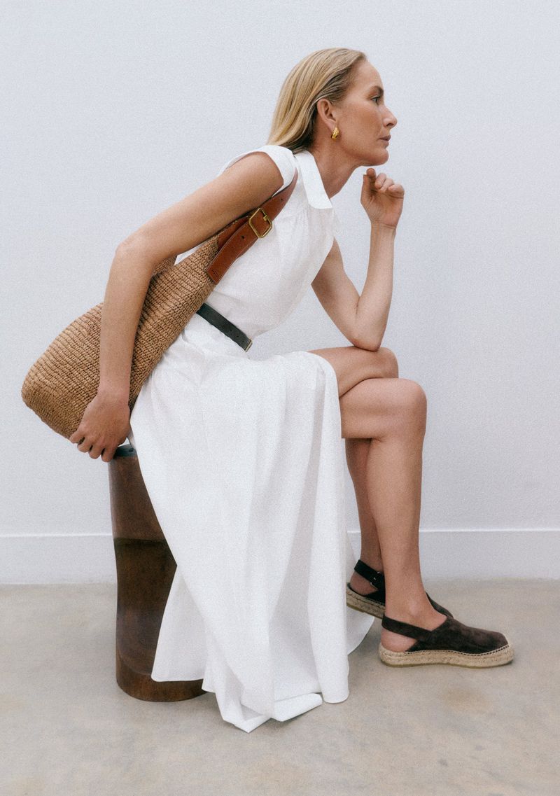 Woman in long white dress sits on a stool, posing in front of a white wall, wearing black espadrilles and a brown shoulder bag.