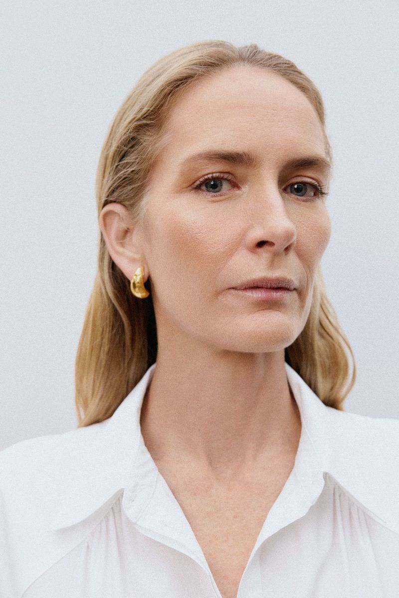 Close-up studio shot of a blonde adult female with gold hoop earrings, fair skin, and a white collared shirt against a light background.