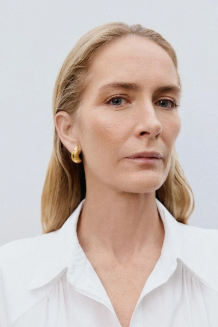 Close-up studio shot of a blonde adult female with gold hoop earrings, fair skin, and a white collared shirt against a light background.
