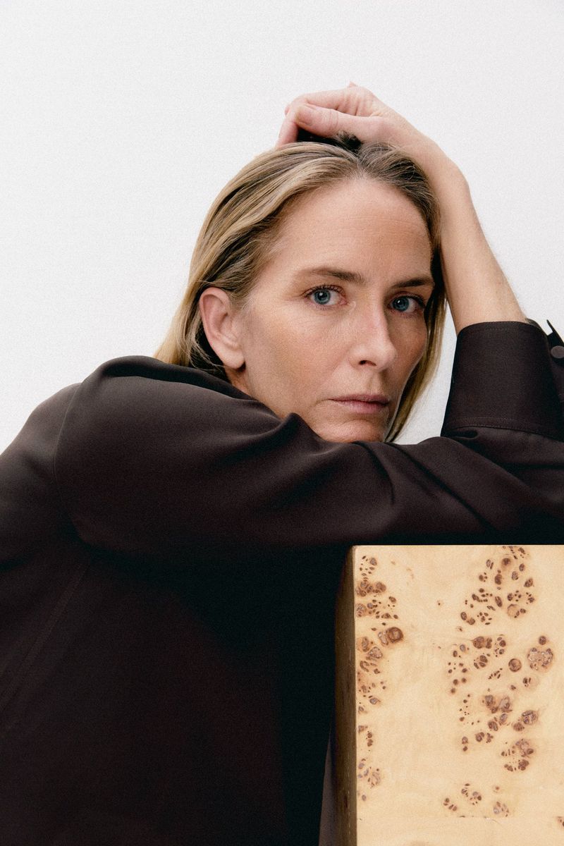 Adult woman in dark brown blouse poses indoors next to small table with decorative pattern in minimalist style portrait with neutral background.