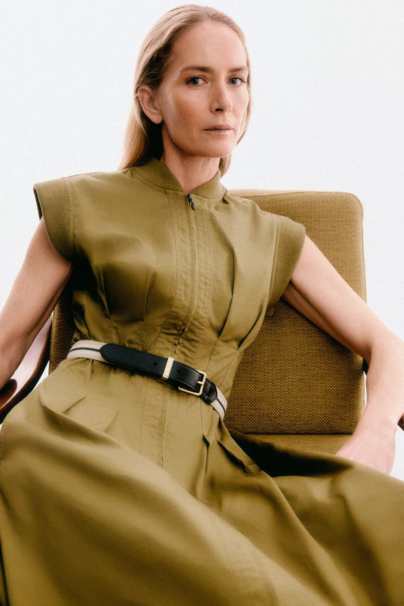 Blonde woman wearing an olive dress with black belt poses in a chair against a white backdrop in a fashion or editorial shot.