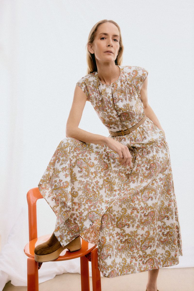 A blonde woman in a patterned blouse and skirt sits on an orange chair in a studio, showcasing a fashion or lifestyle shoot.