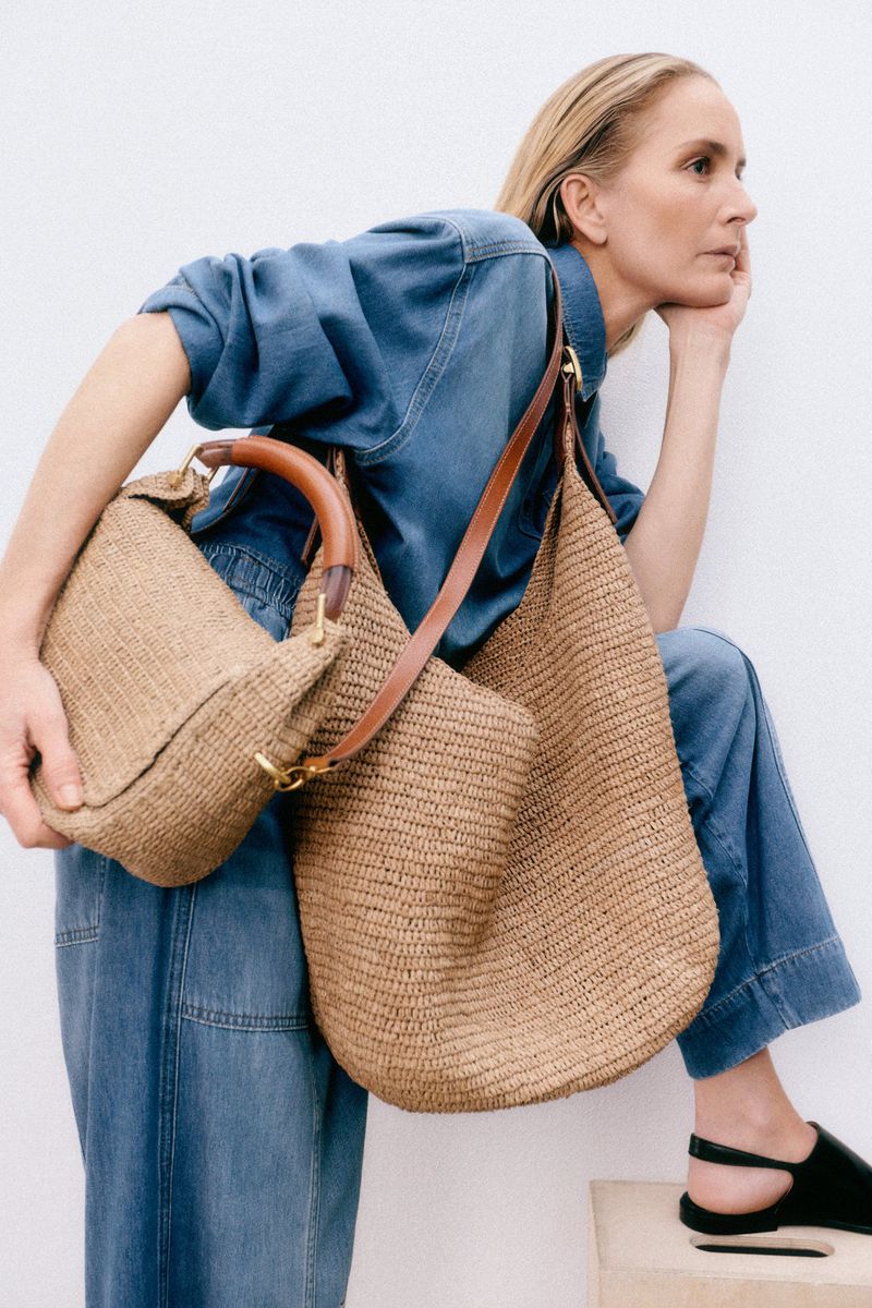 Woman in denim ensemble poses with two straw bags; one is a hobo style, and the other has a handle. Shot against a white background.