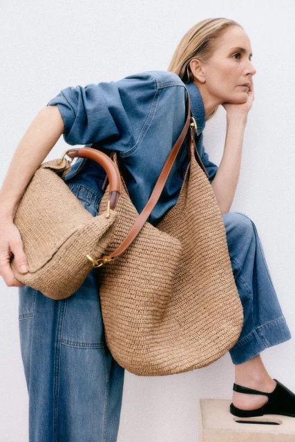 Woman in denim ensemble poses with two straw bags; one is a hobo style, and the other has a handle. Shot against a white background.