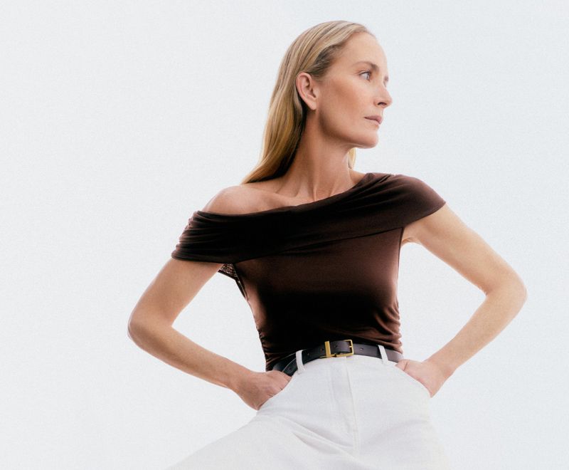 Attractive woman in a brown off-the-shoulder top and white pants poses in a brightly lit photo studio with hands on her hips.