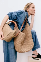 Model in denim shirt and pants holding woven straw bags against a white background