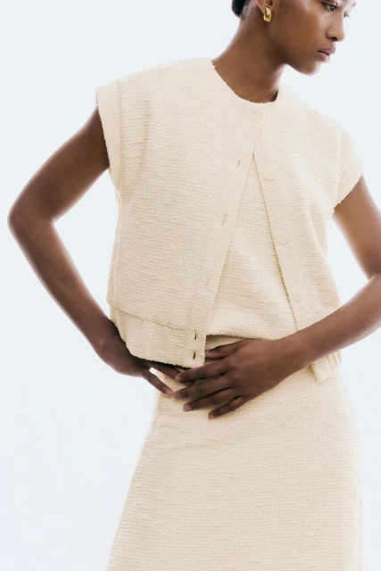 Dark-skinned model poses in a cream-colored tweed vest and skirt ensemble against a white backdrop. She wears gold hoop earrings.