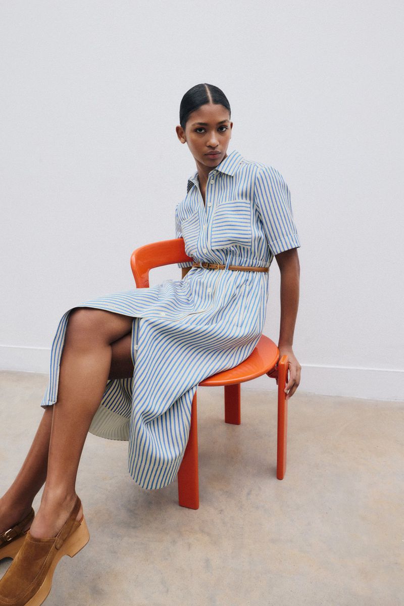 A dark-haired woman is seated on an orange chair, wearing a blue-and-white striped dress with a brown belt, against a white backdrop.