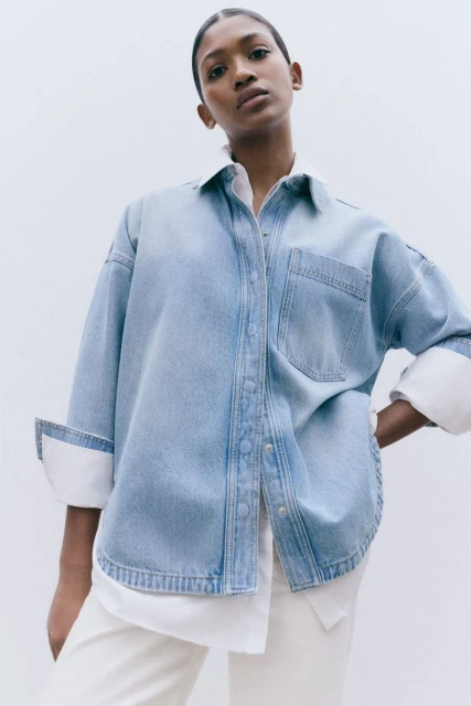 Studio shot of an adult female model wearing a denim shirt with rolled-up sleeves and white pants against a white background.