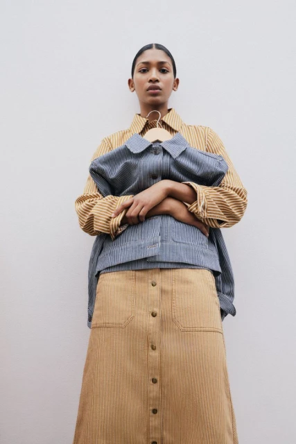 Woman in a striped skirt and shirt holds a striped jacket on a hanger against a white background in a studio-lit, eye-level portrait.