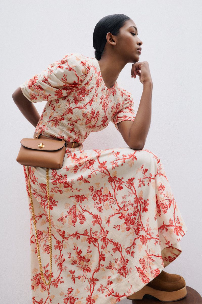 Woman in floral dress and brown clogs sitting indoors in a studio environment, wearing a brown purse and a neutral background.