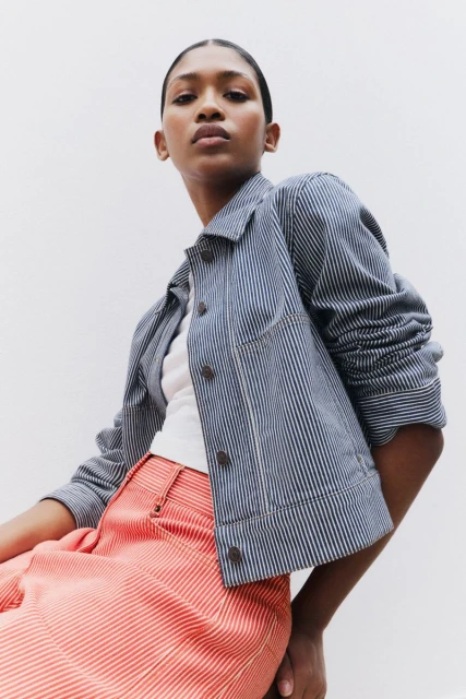 A stylish portrait of a young adult woman, wearing a striped jacket and pants, shot at a low angle against a white background.