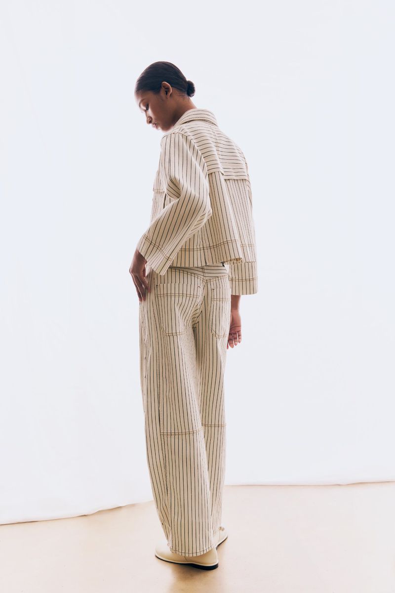 Fashionable adult female posing in a photo studio. She is wearing a vertically striped tan jacket and pants with neutral shoes.