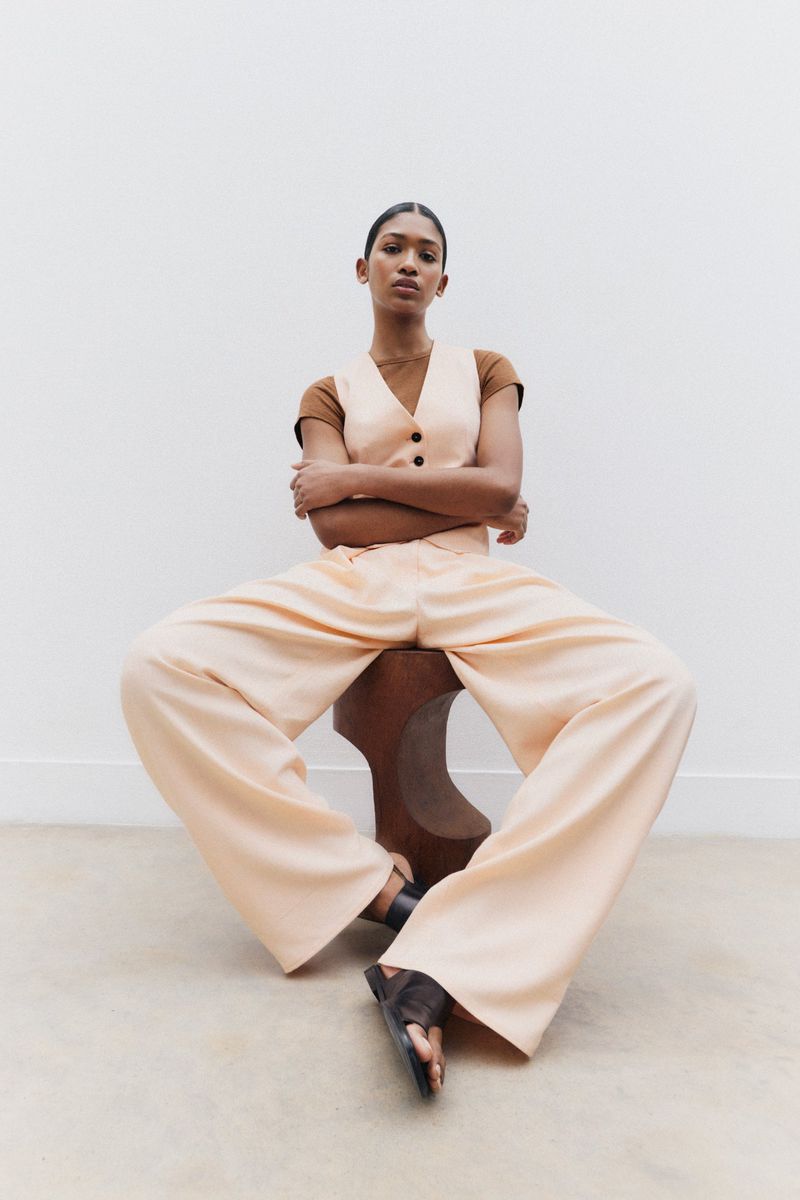 A stylish woman in a peach linen suit sits on a wooden stool against a neutral wall in a studio. Minimalist fashion shot.