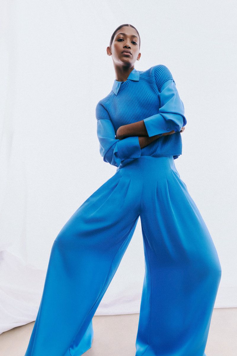 A woman in a blue shirt and flowing pants stands against a white backdrop in a low-angle fashion shot with studio lighting.