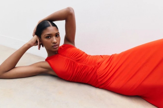 Model wearing a vibrant orange ruched dress lying down against a plain white studio background for ME+EM