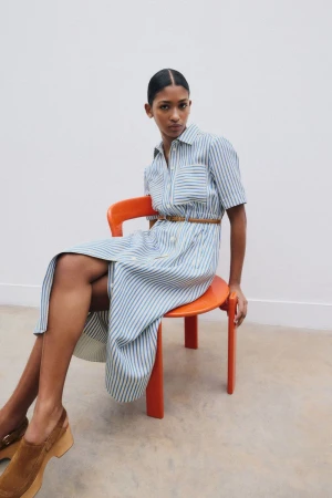 Model wearing a blue striped shirt dress sitting on a bright orange chair against a white wall.