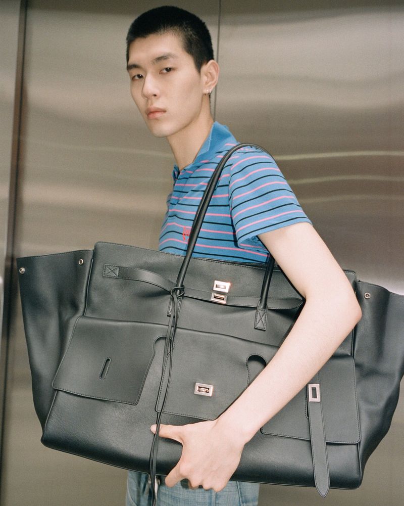 Stylish Asian man in a blue striped polo shirt carries a large black leather tote bag in a modern, neutral indoor environment.