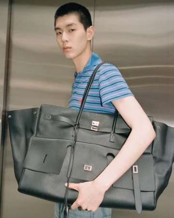 Stylish Asian man in a blue striped polo shirt carries a large black leather tote bag in a modern, neutral indoor environment.
