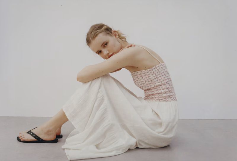 Woman in white skirt and patterned top sits on the floor in a fashion pose, angled view, studio setting, natural light.