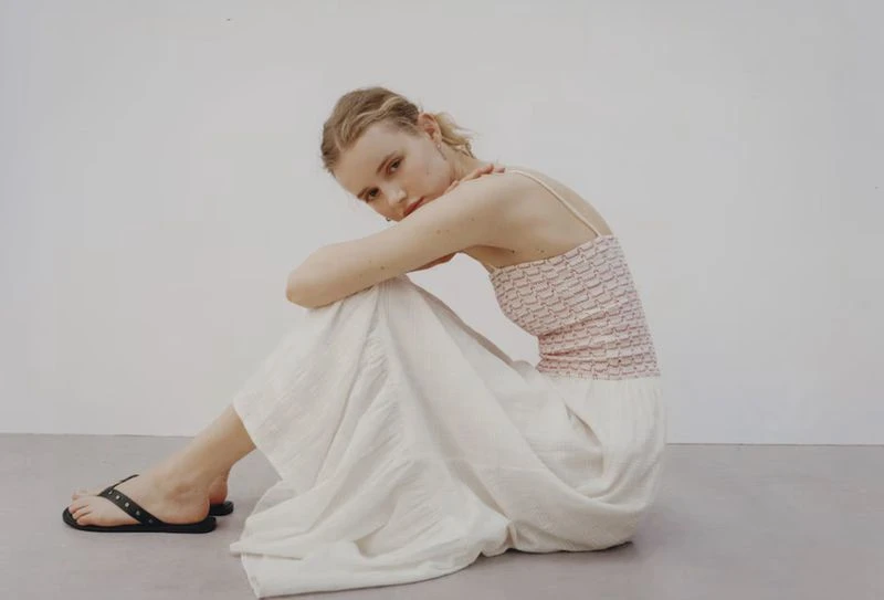 Woman in white skirt and patterned top sits on the floor in a fashion pose, angled view, studio setting, natural light.