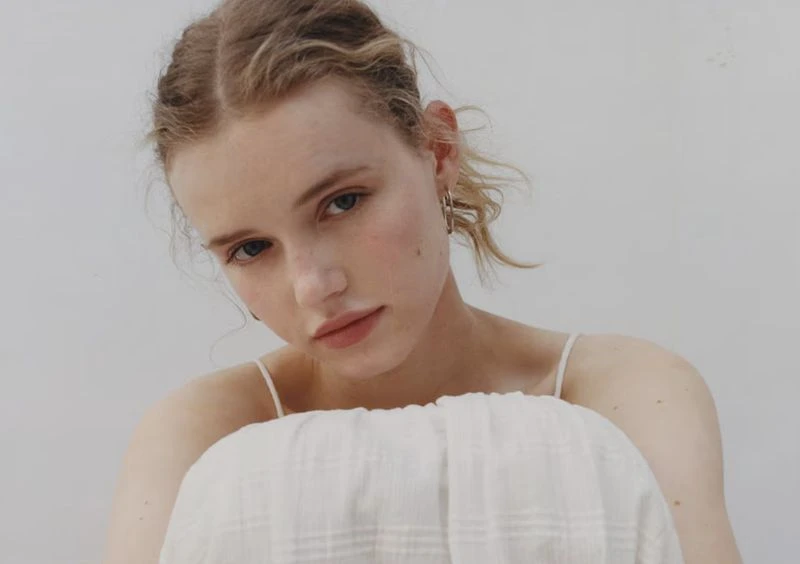 Close-up portrait of a young adult woman with blonde hair, fair skin, light make-up, wearing a white top, against a bright, plain background.