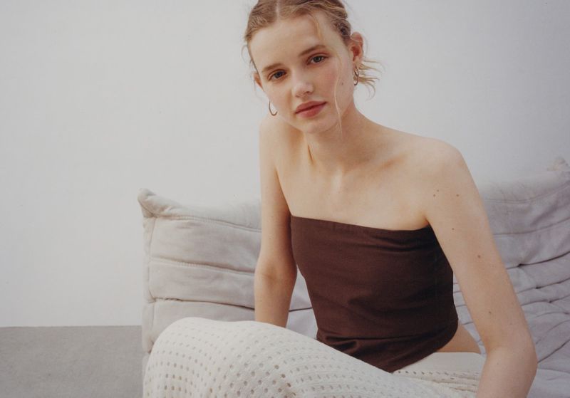 Model wearing a brown strapless top and cream patterned skirt seated on a light grey sofa against a white wall.