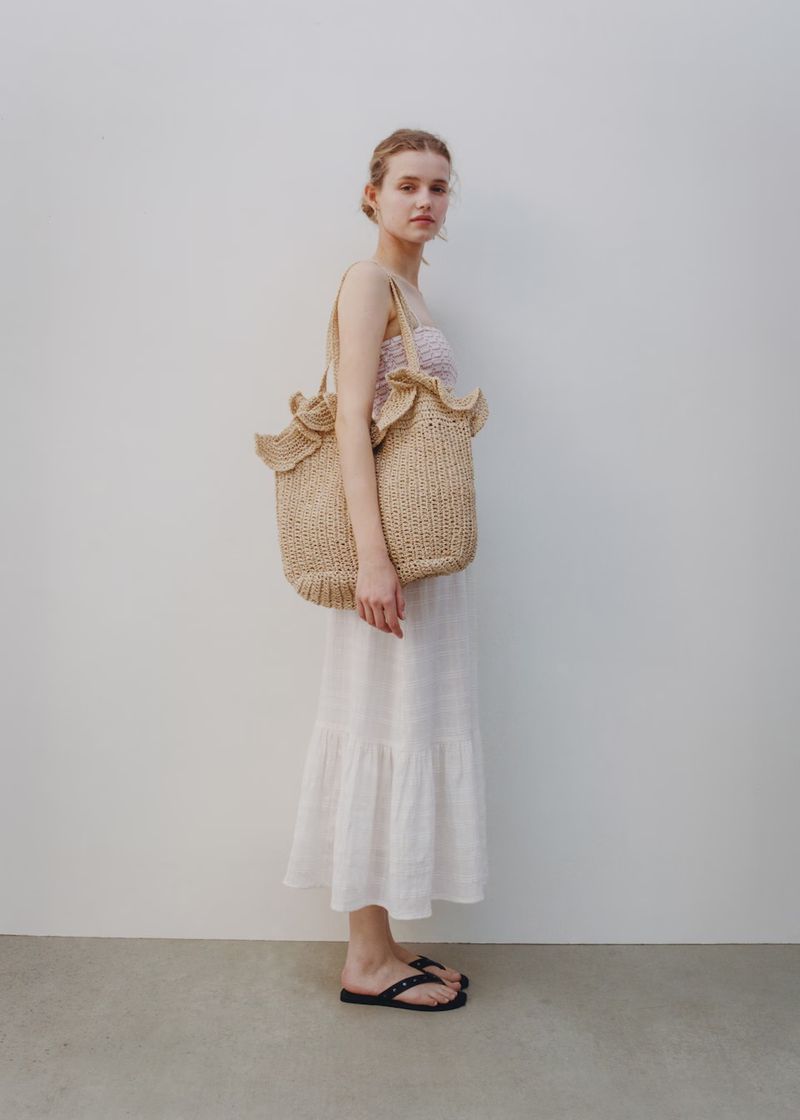 Woman in white dress and black sandals poses with a straw tote bag against a neutral background in a full shot.