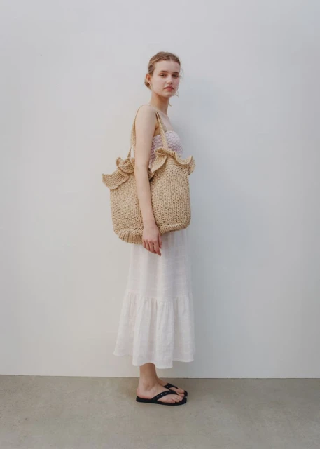 Woman in white dress and black sandals poses with a straw tote bag against a neutral background in a full shot.