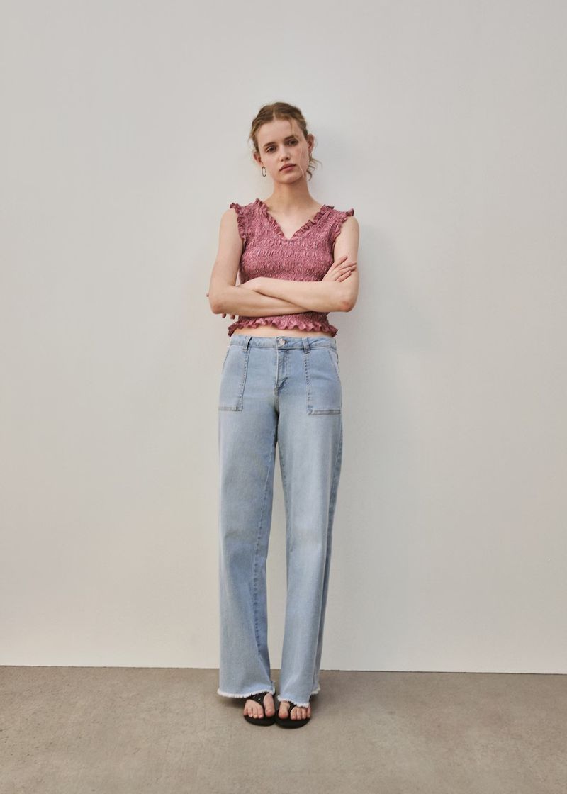 A woman stands in a photo studio wearing a pink sleeveless top and wide-leg denim jeans, posing with arms crossed in a full shot.