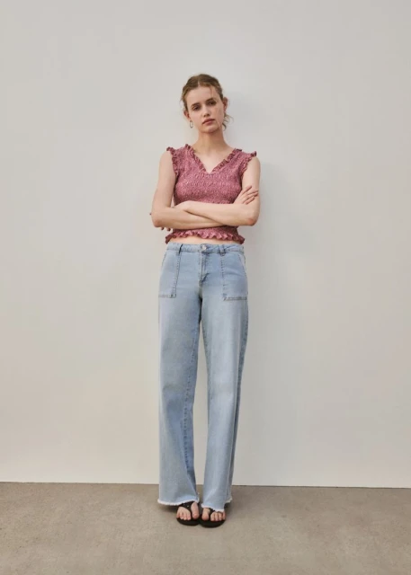 A woman stands in a photo studio wearing a pink sleeveless top and wide-leg denim jeans, posing with arms crossed in a full shot.