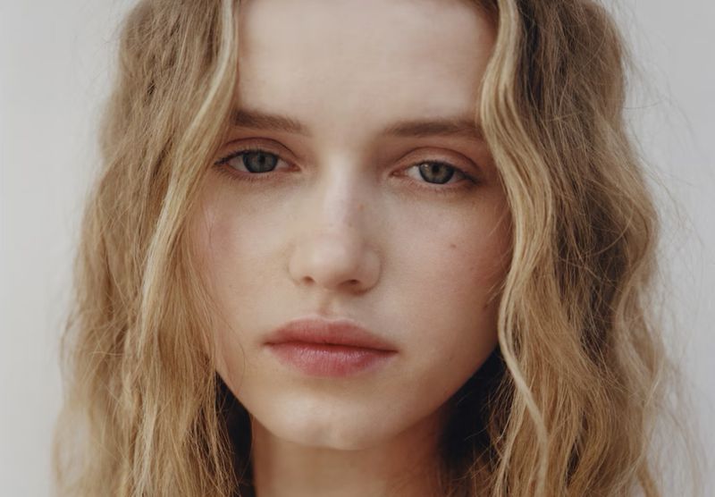 Close-up portrait of a young female model with blonde wavy hair and neutral expression against a plain white background.