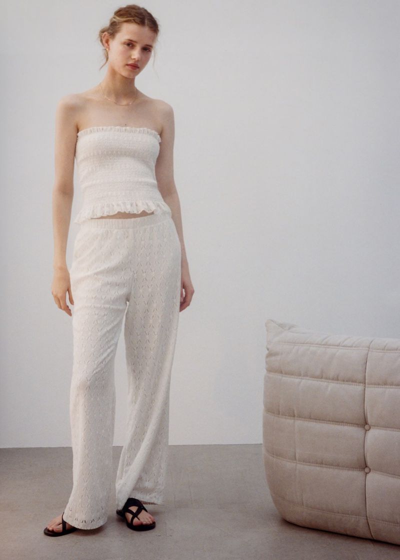 A young woman in a white strapless top and matching wide-leg pants stands indoors near a beige cushion and gray flooring.