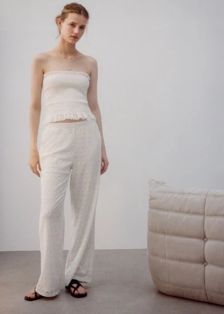 A young woman in a white strapless top and matching wide-leg pants stands indoors near a beige cushion and gray flooring.