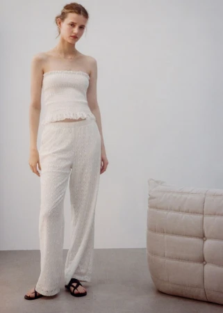 A young woman in a white strapless top and matching wide-leg pants stands indoors near a beige cushion and gray flooring.