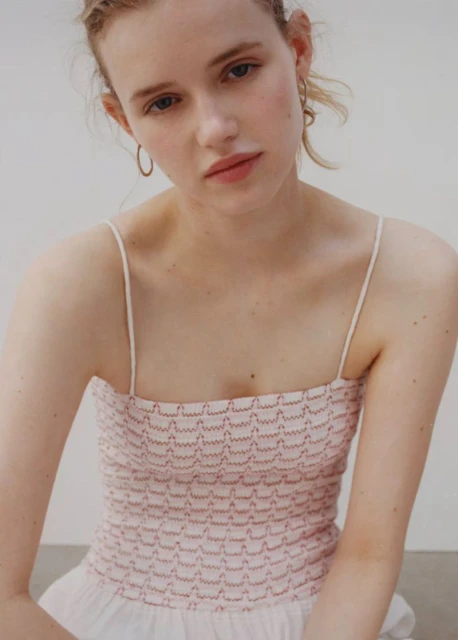 Portrait of a young woman with fair skin and blonde hair, wearing a pink and white top, in a neutral-toned indoor setting. Soft, diffused lighting.