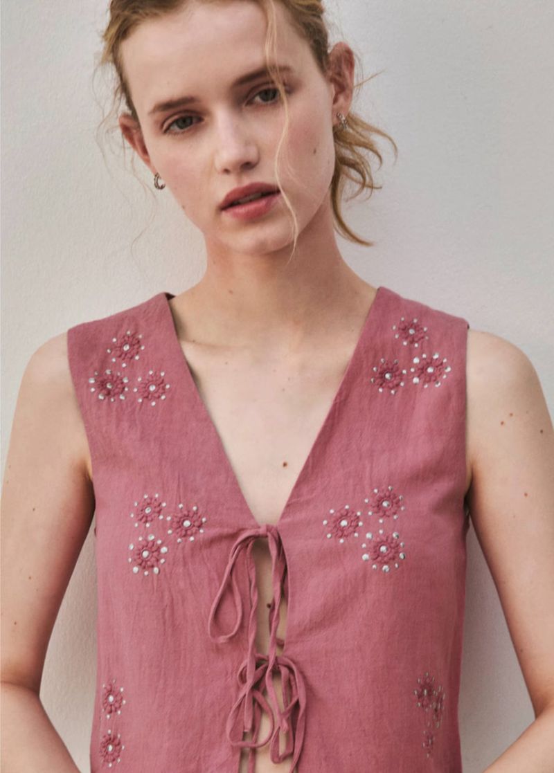Young woman wearing a pink sleeveless top with floral embroidery, posing in a brightly lit, neutral-toned indoor setting. Close-up.