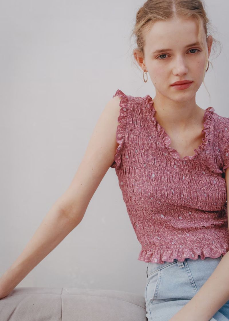 Young adult woman poses indoors wearing a smocked pink top and light blue jeans, looking directly at the camera with a neutral expression.
