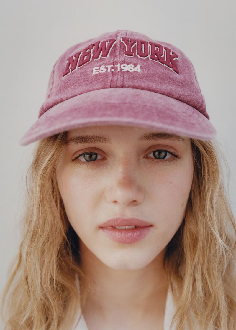 Close-up portrait of a blonde woman wearing a pink baseball cap embroidered with 'New York EST. 1984,' set against a neutral background.