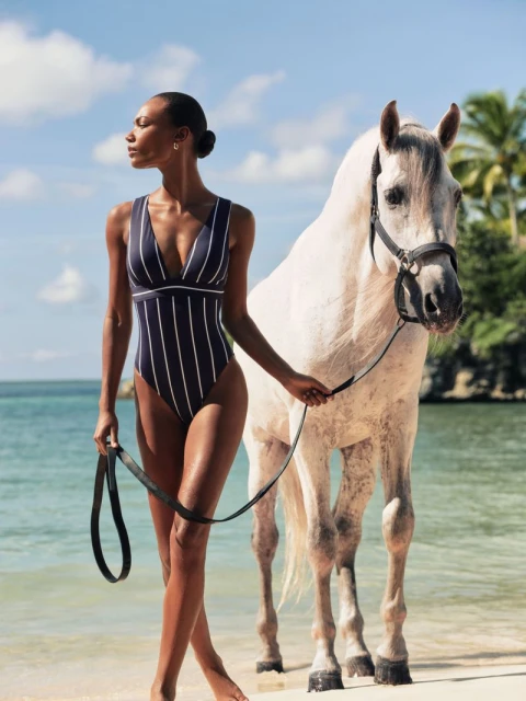 Woman in striped swimsuit poses with a white horse on the beach with ocean and sky in the background on a sunny day.