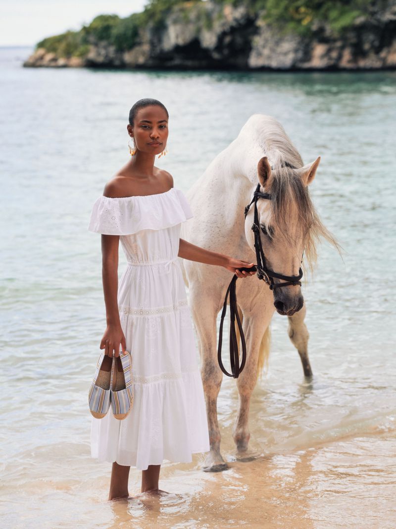 Model in white off-shoulder dress holding espadrilles standing with a white horse in shallow water at the beach