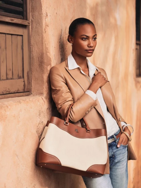 Adult female fashion model in beige blazer stands with a tote bag against a textured stucco wall with a window, casual, neutral palette.