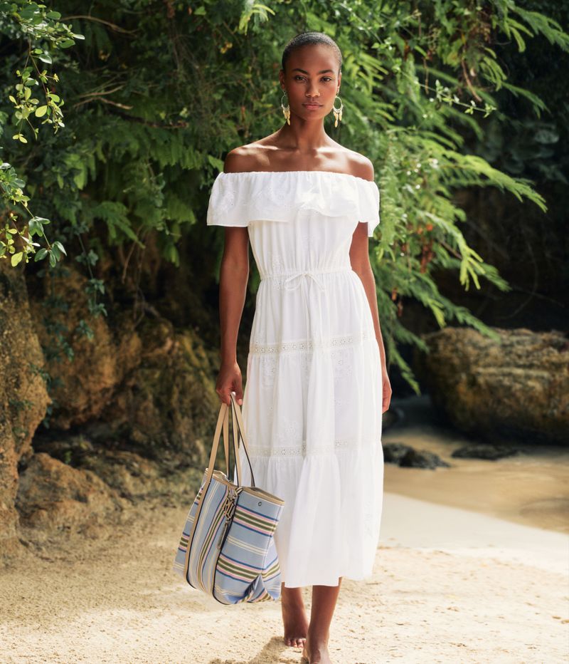 Woman in white dress on beach carries striped tote bag, accessorized with earrings. Verdant foliage and sand frame the scene.